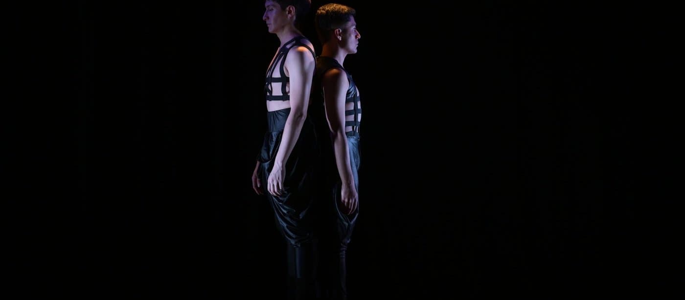 Two dancers standing back to back under a single spotlight on a dark stage