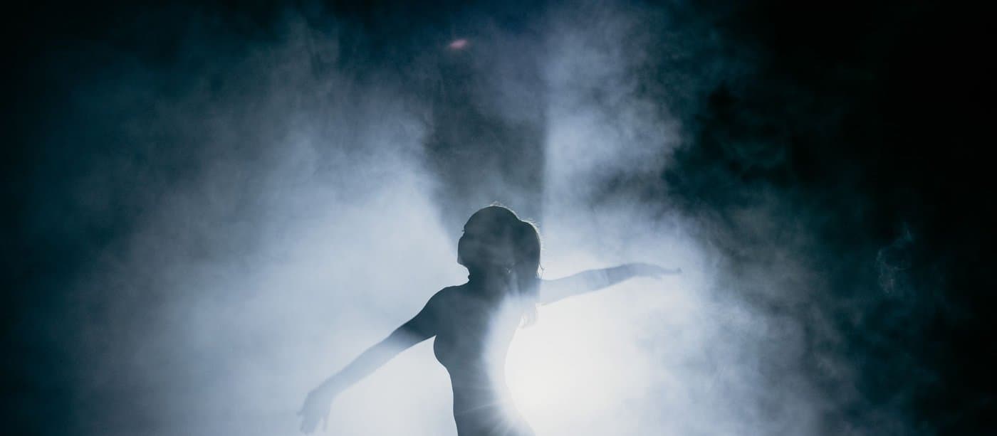A dancer silhouetted against a spotlight and stage smoke, arms extended mid-move